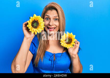 Femme hispanique d'âge moyen tenant des tournesols jaunes expression sans indice et confuse. Doute concept. Banque D'Images