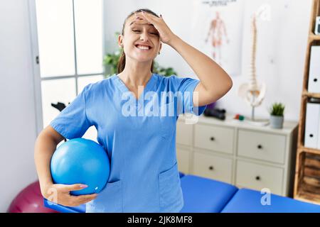 Jeune physiothérapeute femme tenant le ballon pilates à la clinique médicale stressée et frustrée avec la main sur la tête, surpris et fâché visage Banque D'Images