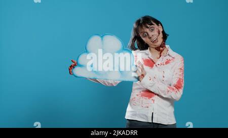 Femme zombie créepy tenant un art de nuage de carton tout en se tenant sur fond bleu. Monstre dangereux effrayant et terrifiant, avec des blessures sanglantes et profondes et des cicatrices ayant un nuage bleu de papier. Prise de vue en studio Banque D'Images