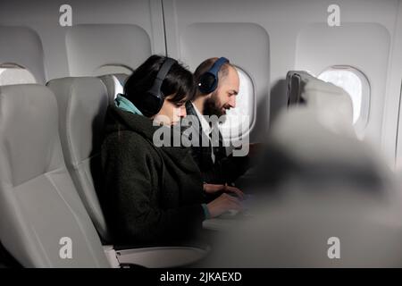 Jeunes voyageurs utilisant un ordinateur portable et un smartphone en avion, attendant d'arriver aux vacances. Deux passagers travaillant sur des appareils et voyageant avec une compagnie aérienne internationale. Banque D'Images