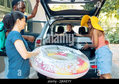 Famille mixte et amis chargeant des sacs dans le coffre, préparant des bagages et une valise en automobile. Voyager en bord de mer avec des gens divers, mettre des bagages et gonflables dans le véhicule, partir en vacances. Banque D'Images