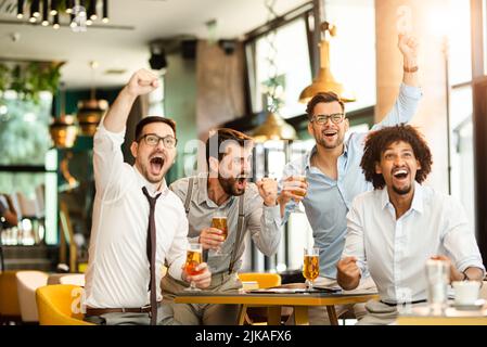 Quatre jeunes hommes vêtés de vêtements décontractés applaudissent au football et tiennent des verres de bière tout en étant assis au pub Banque D'Images