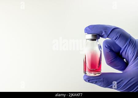 Liquide rouge (vitamine B12) dans une ampoule en verre avec un bouchon dans la main dans un gant médical bleu sur fond blanc. Beaucoup d'espace vide Banque D'Images