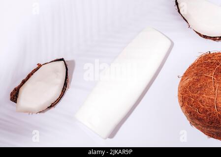 Bouteille cosmétique blanche et noix de coco fraîches sur fond blanc dans l'ombre des feuilles de palmier. Cosmétiques naturels biologiques à l'huile de coco. Vue de dessus, plan d'appartement Banque D'Images