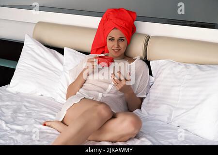 Portrait d'une jeune femme regardant l'appareil photo. Utilise le téléphone pour lire les actualités, faire défiler les médias sociaux tout en étant allongé avec une serviette rouge sur la tête dans la chambre Banque D'Images