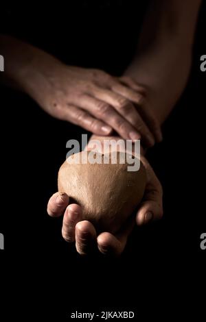 Mains de récolte personne méconnaissable tenant l'argile en forme de coeur pour faire de la vaisselle sur fond noir dans un studio moderne avec une faible lumière Banque D'Images