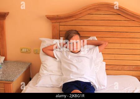 Un garçon se trouve sur un lit avec des draps blancs dans une chambre d'hôtel. L'enfant dort. Adolescent au lit à la maison Banque D'Images
