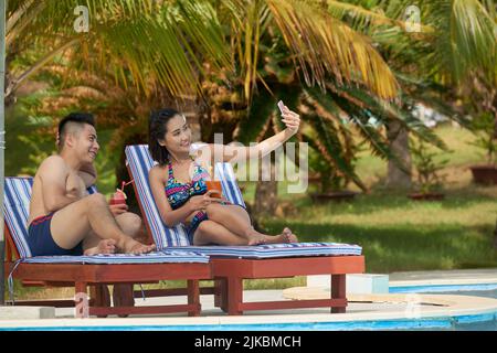 Un jeune couple heureux prend un bain de soleil au bord de la piscine et prend des selfies Banque D'Images