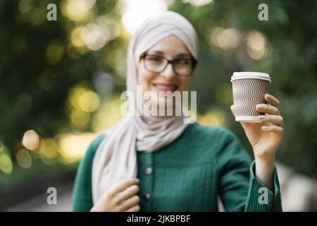 Concentrez-vous sur une tasse de café de la femme musulmane souriante attrayante dans le hijab debout sur fond vert du parc de la ville. Portrait d'une jeune femme arabe avec lunettes et boisson chaude à l'extérieur. Banque D'Images