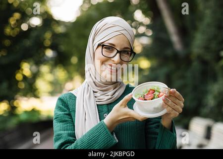 Joyeuse femme musulmane manger un déjeuner sain à l'extérieur. Femme en vêtements décontractés avec fourchette à la main et salade de légumes dans un bol, sur fond de parc vert, concept de nourriture de régime. Banque D'Images