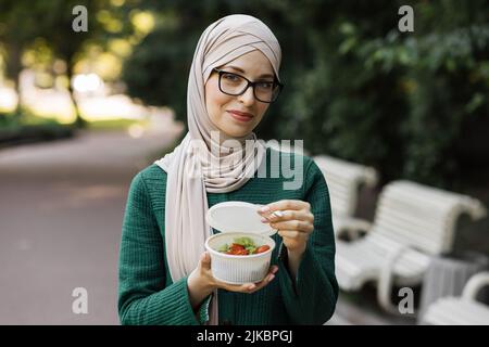 Joyeuse femme musulmane manger un déjeuner sain à l'extérieur. Femme en vêtements décontractés avec fourchette à la main et salade de légumes dans un bol, sur fond de parc vert, concept de nourriture de régime. Banque D'Images