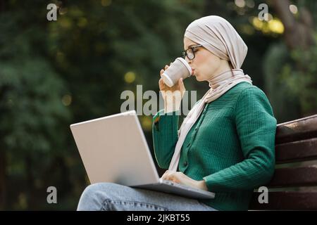 Une femme d'affaires musulmane souriante qui boit du café pendant une pause, assise sur un banc et travaillant sur un ordinateur portable. Belle fille souriante de l'islam appréciant le temps pendant la pause-café. Banque D'Images