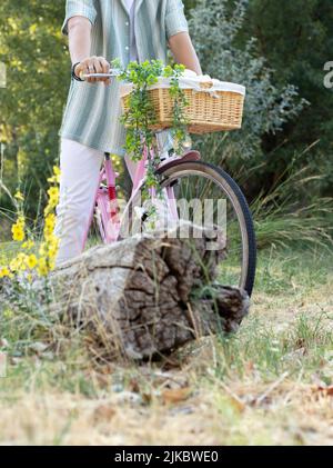 Vue verticale courte d'un jeune homme méconnu sur une bicyclette rose vintage avec panier et plantes et une bûche devant Banque D'Images