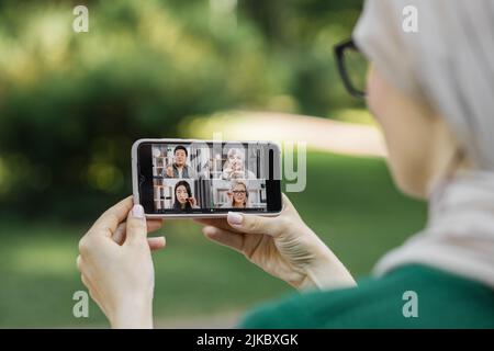 Gros plan sur de jolies jeunes musulmanes en hijab beige, en appréciant du temps dans le parc d'été, tout en parlant avec quatre collègues multi-ethniques à l'aide d'un smartphone. Banque D'Images