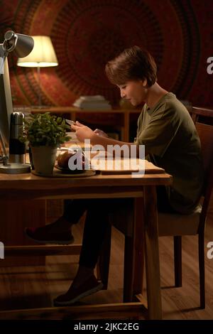 Vue latérale d'un étudiant utilisant une tablette numérique tout en faisant ses devoirs à la lumière d'une lampe Banque D'Images