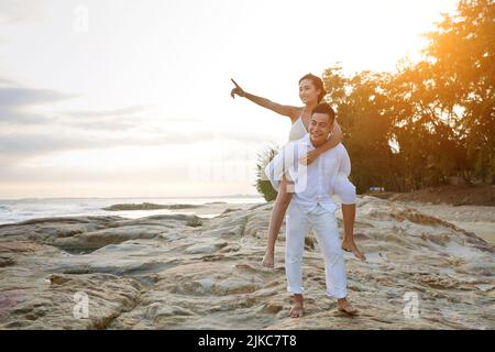 Homme asiatique gai portant un pantalon et une chemise blancs et regardant loin avec un sourire crasseux tout en offrant une promenade à dos de porc à sa jolie petite amie, le rivage de sable de mer et le coucher de soleil pittoresque sur fond Banque D'Images