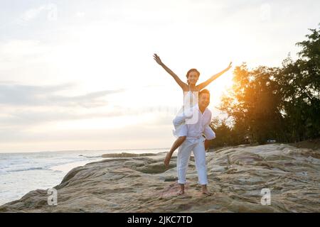 Couple amoureux en vacances dans un complexe exotique : homme asiatique joyeux qui donne un voyage en pigeyback à sa belle petite amie aux bras étirés, paysage marin pittoresque sur fond Banque D'Images