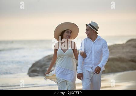 Joyeux couple asiatique portant des vêtements blancs discutant avec animation les uns avec les autres tout en marchant le long de la mer à un coucher de soleil à couper le souffle Banque D'Images