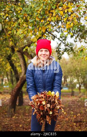 Défocus jeune fille souriante tenant des feuilles sèches. Un enfant volontaire ratisse et saisit une petite pile de feuilles jaunes tombées dans le parc d'automne. Nettoyage de la Banque D'Images