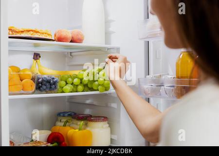 Une femme prend à la main, saisit ou ramasse des raisins verts dans une étagère de réfrigérateur ouverte ou un tiroir de réfrigérateur plein de fruits, légumes, banane, pêches, yaourt. Alimentation saine alimentation, concept de mode de vie Banque D'Images