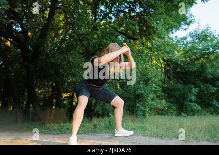 modèle de forme physique de fille mince dans un survêtement noir exécute des squats tenant ses mains devant elle Banque D'Images