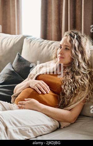 Femme enceinte aux cheveux longs blonds ondulés gardant les mains sur son ventre tout en se relaxant sur un canapé doux et en regardant la télévision à votre guise Banque D'Images