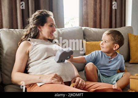 Bonne jeune femme blonde enceinte regardant son mignon petit fils mettant jouet animal sur le ventre de sa mère assis devant lui Banque D'Images