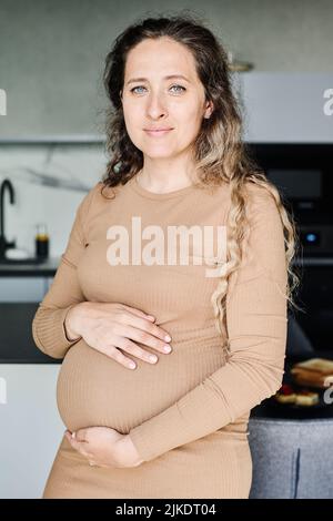 Jeune jolie femme enceinte en robe brune décontractée regardant l'appareil photo tout en se tenant dans la cuisine et en touchant son ventre Banque D'Images