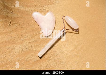 Pose plate d'un rouleau de jade en pierre rose et d'un masseur gua-sha, isolé sur un fond de sable doré avec espace de copie publicitaire Banque D'Images