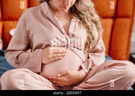 Jeune femme enceinte en casque d'intérieur rose touchant son ventre avec le futur bébé tout en étant assise sur le lit et en se mettant prêt à dormir Banque D'Images