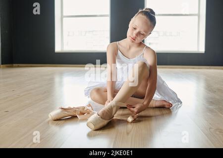 Jeune blonde ballerine en blanc tutu et beige leggins attacher ses chaussures pointe avant la répétition tout en étant assis sur le sol du studio Banque D'Images