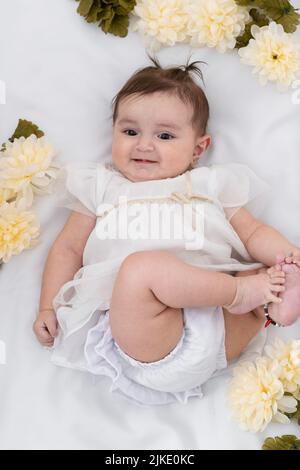 photo aérienne d'un beau bébé latina, allongé sur un drap blanc, souriant très heureux tout en tenant ses pieds, entouré de fleurs et habillé dans un Banque D'Images