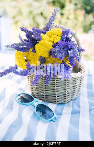 bouquet d'été d'yarrow jaune et de lavande violette dans un panier avec des lunettes de vue brillantes sur une couverture de pique-nique rayée avec la vibe d'été Banque D'Images
