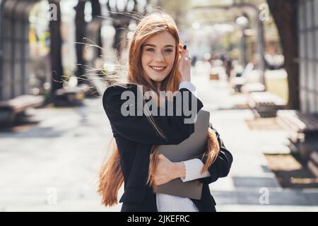 Femme d'affaires inconsidérée de race blanche tenant son ordinateur portable pendant qu'elle va travailler dans le parc urbain. Jeune femme aux cheveux rouges dans le parc. Femme avec un ordinateur portable essayant Banque D'Images