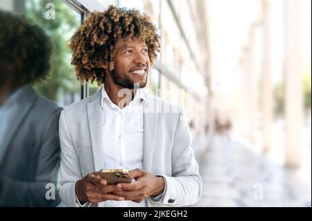 Positif réussi brésilien ou africain américain avec des cheveux bouclés, élégamment habillé, debout à l'extérieur près du centre d'affaires, en utilisant le téléphone mobile, regardant sur le côté, rêvant, sourire Banque D'Images