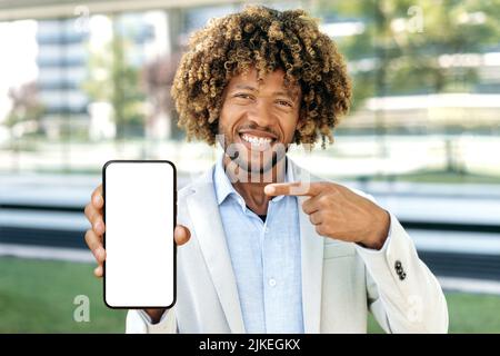 Beau et heureux afro-américain mauriquement cheveux homme, se tient à l'extérieur, montre son smartphone avec écran noir de maquette blanc, pointe le doigt sur lui, espace pour la publicité, la présentation, regarde l'appareil photo, sourire Banque D'Images