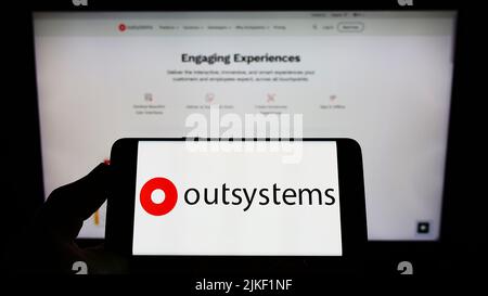 Personne tenant un téléphone portable avec le logo de la société de logiciel à code faible OutSystems à l'écran en face de la page Web d'affaires. Mise au point sur l'affichage du téléphone. Banque D'Images