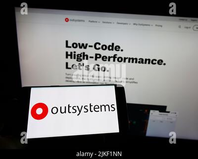 Personne tenant le téléphone portable avec le logo de la société de logiciel à code faible OutSystems à l'écran devant la page Web d'affaires. Mise au point sur l'affichage du téléphone. Banque D'Images