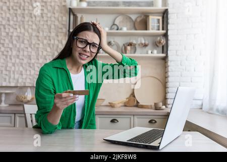 Portrait d'une femme insatisfaite et déçue à la maison, brunette essayant de faire un achat en magasin en ligne, a été rejeté, tenant la carte de crédit de banque et regardant l'appareil photo malheureusement Banque D'Images