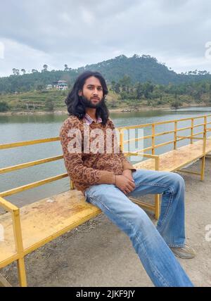 Un jeune indien à l'aspect agréable avec des cheveux longs et une barbe regardant l'appareil photo tout en étant assis sur un banc à côté du lac Banque D'Images