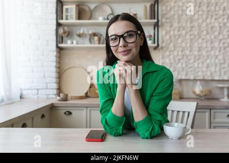 Jeune belle femme en lunettes et une chemise verte décontractée, à la maison dans la cuisine, regarde la webcam, appel vidéo écoute l'interlocuteur, vue de la webcam Banque D'Images