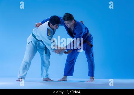 Portrait dynamique d'athlètes professionnels judo s'entraîner isolé sur fond bleu studio dans la lumière du néon. Exercice sur la roue d'épaule Banque D'Images