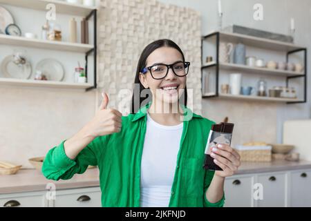 Jeune belle femme adolescente tenant la barre de chocolat dans les mains dans des verres et une chemise verte, regardant l'appareil photo sourire, recommandant de tenir le pouce vers le haut Banque D'Images