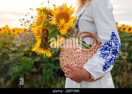 Gros plan du sac de paille rempli de tournesol. Femme tenant un sac à main d'été avec bouquet de fleurs dans le champ au coucher du soleil Banque D'Images