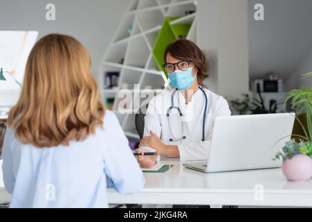 Femme médecin portant un masque facial pendant qu'elle travaille au bureau et qu'elle écoute le patient. Banque D'Images