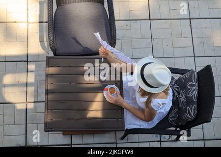 Une femme solitaire avec un chapeau se trouve dans un café extérieur. Seule femme tenant un ventilateur de main et se refroidissant elle-même un jour d'été. Gros plan, vue de dessus Banque D'Images