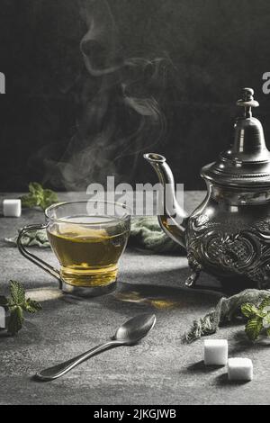 Théière marocaine traditionnelle avec une tasse de thé à la vapeur dans une atmosphère noire fumée. Format vertical. Banque D'Images