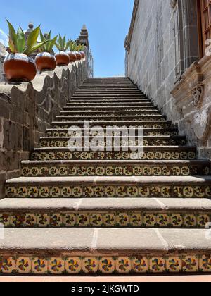 Un escalier extérieur mène au ciel bleu, avec des carreaux décoratifs, des pots à motifs et un passage le long d'un mur adjacent. Banque D'Images