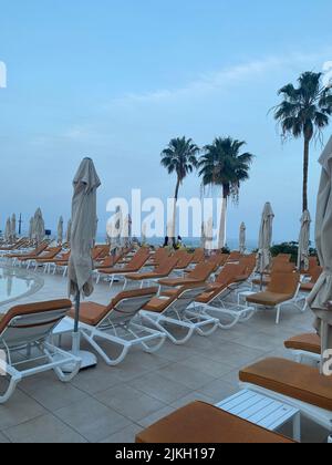 A vertical shot of sun loungers with umbrellas by the pool with palm trees in the background Stock Photo