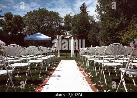 An outdoor wedding venue with an aisle with floral petals and white chairs in a park Stock Photo